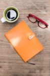 Blank Orange Leather Diary,glasses And A Cup Of Coffee On Wooden Desk Stock Photo
