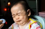 Close Up Of Unhappy Little Seven Months Old Son In See Through Plastic Bib Screaming And Crying In Chair For Babies After Mom Made Him Eat Avocado And Banana Mix.unhappy Baby's Face. Asian Infant Cry Stock Photo