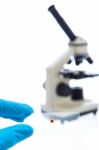 Hand In Blue Latex Glove Holds Sample Glass Plate With Blood Sample Drop And A Microscope On White Background Stock Photo