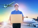 Delivery Man Holding Card Box Toothy Smiling Face Against Shipping Port And Cargo Plane Flying Background Stock Photo
