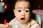 Close Up Of Happy Little Seven Months Old Son In See Through Plastic Bib Eating In Chair For Babies After Mom Made Him Eat Avocado And Banana Mix.happy Baby's Face. Asian Infant Open Mouth Stock Photo
