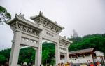 Tourists At The Gate To Po Lin Monastery With Tian Tan Buddha Statue Up On The Hill In Background, Ngong Ping Village, Lantau Island, Hong Kong Stock Photo