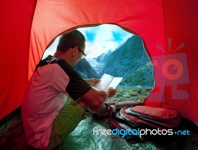 Camping Man Reading Traveling Guide Book In Camp Tent Against Beautiful Scenic Of Glacier Mountain Stock Photo