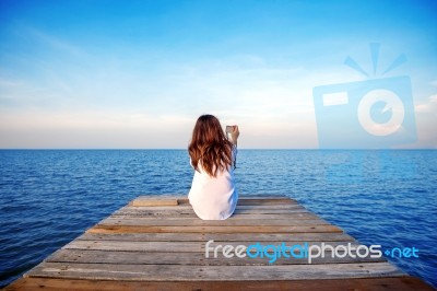 Girl Sitting Alone And Hand Holding Camera On A The Wooden Bridge On The Sea Stock Photo