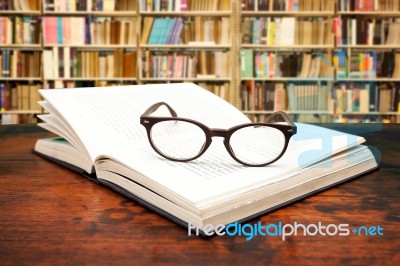 Open Book With Glasses On The Desk Against Library Stock Photo