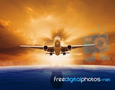 Passenger Jet Plane Flying Over Beautiful Sea Level With Sun Set Sky Above And Urban Skyline Behind Stock Photo
