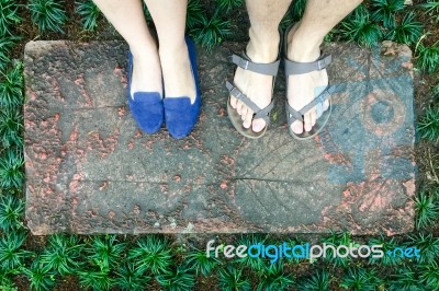 Top View Feet Of Male And Female Couple Lover Standing On Stone Floor Among Little Green Grass Garden. Hipster Style Stock Photo