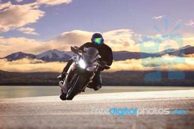 Young Man Riding Big Bike Motorcycle Against Sharp Curve Of Asphalt High Ways Road With Rural Lake Scene Use For Male Adventure Activities And Motor Sport Hobby On Holiday Vacation Stock Photo