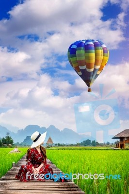 Young Woman Look At Balloon And Sitting On Wooden Path With Green Rice Field In Vang Vieng, Laos Stock Photo