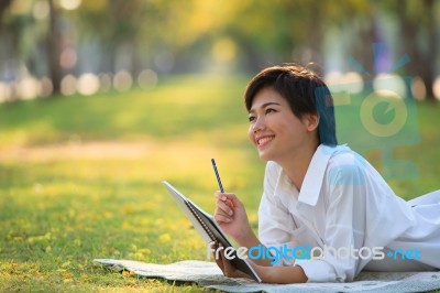 Young Woman Lying On Green Grass Park With Pencil And Note Book Stock Photo