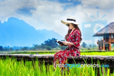 Young Woman Reading A Book And Sitting On Wooden Path With Green Rice Field In Vang Vieng, Laos Stock Photo