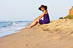 Cute White Girl In Hat And Purple Dress Sitting On Sand Stock Photo