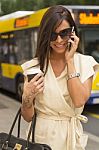 Fashionable Young Brunette Laughs Into Phone, Bus In Background Stock Photo