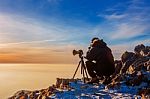 Professional Photographer Takes Photos With Camera On Tripod On Rocky Peak At Sunset Stock Photo